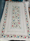 A square white embroidered table runner or decorative textile laid flat, featuring a central rectangular panel with green vines and small red, blue, and purple flowers, surrounded by a matching floral border.