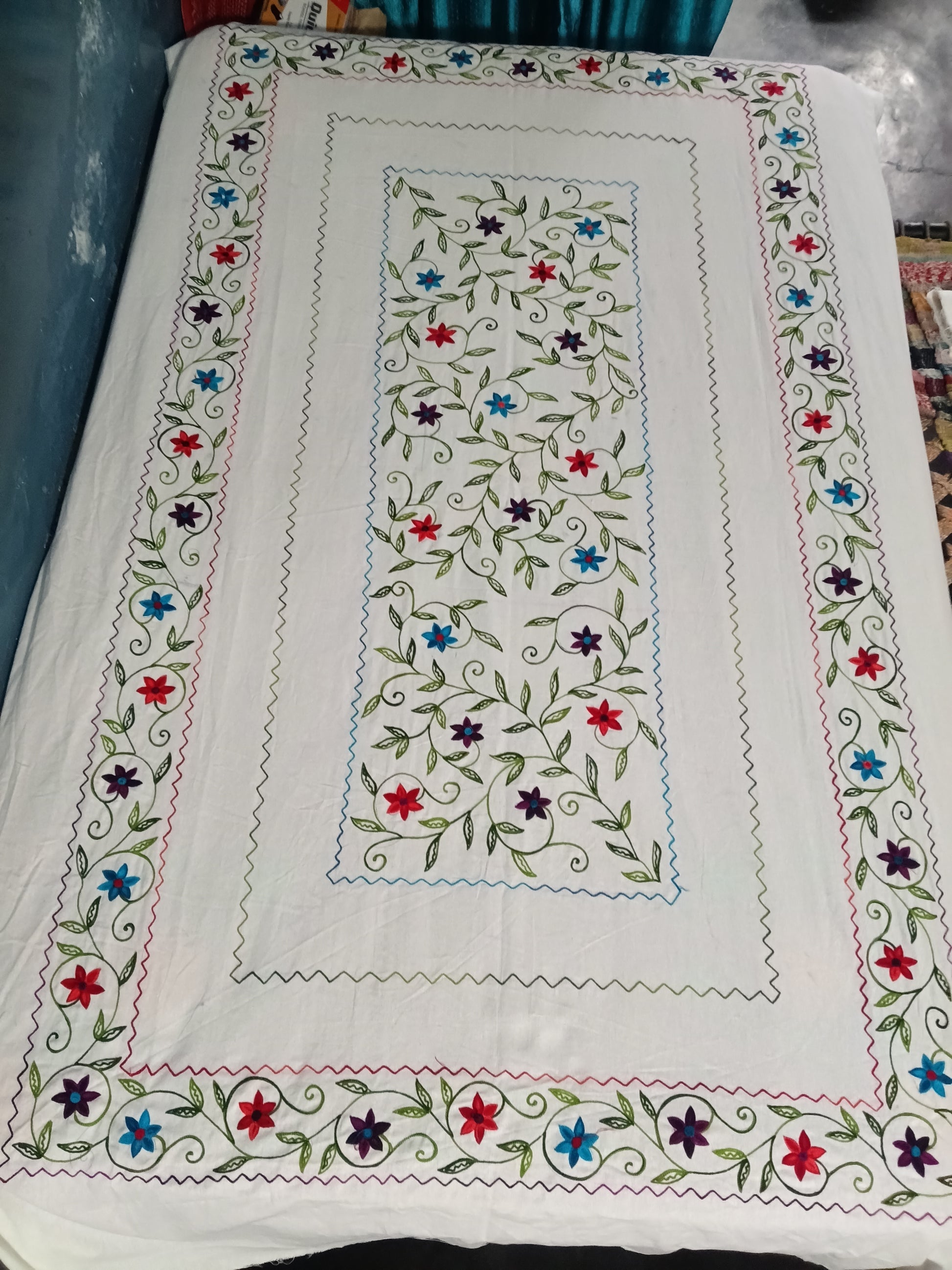 A square white embroidered table runner or decorative textile laid flat, featuring a central rectangular panel with green vines and small red, blue, and purple flowers, surrounded by a matching floral border.