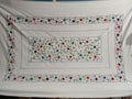 A white embroidered tablecloth featuring a central rectangular floral motif and a wide decorative border with blue, red, and purple flowers, green leaves, and swirling vine details.