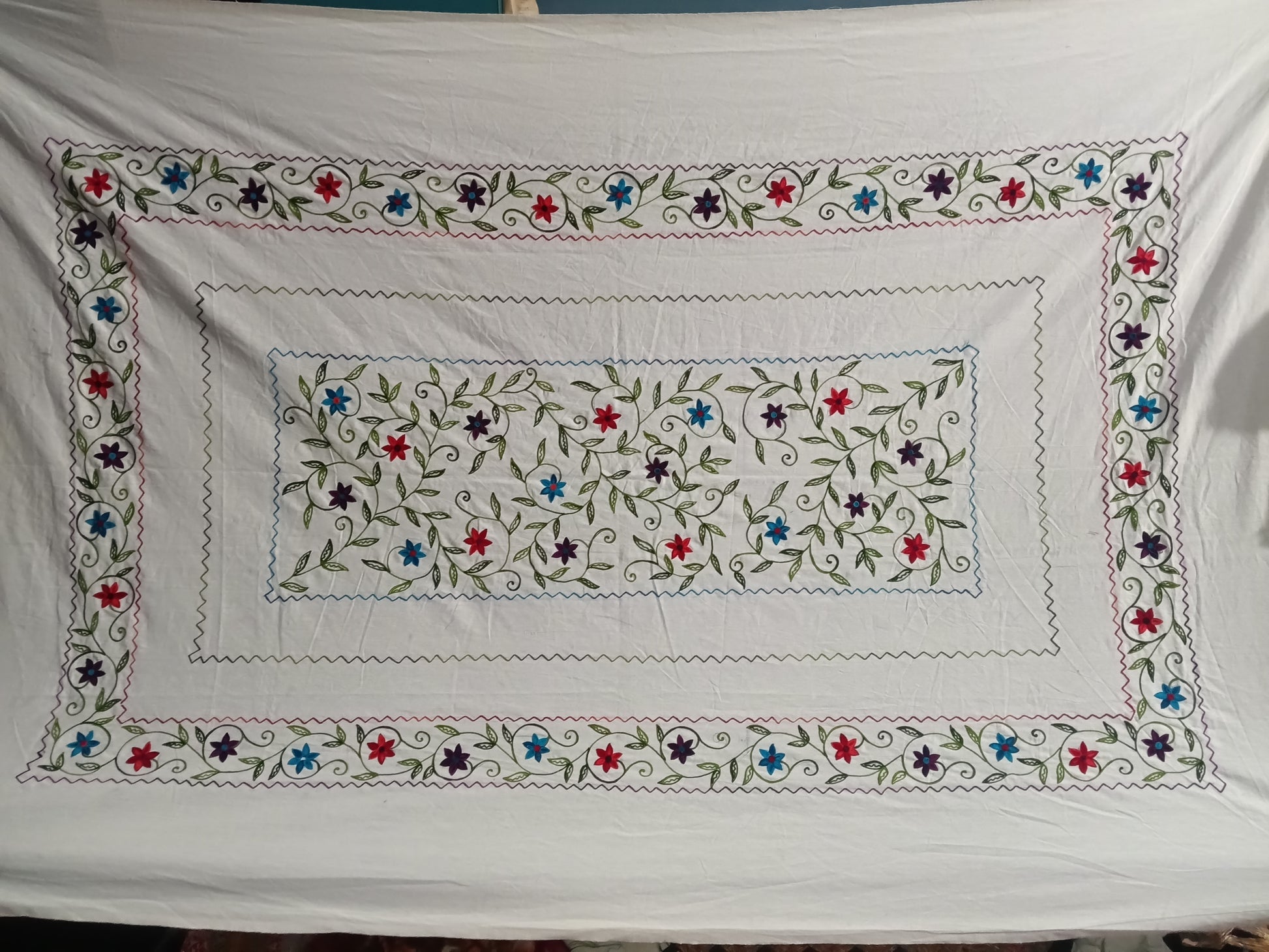 A white embroidered tablecloth featuring a central rectangular floral motif and a wide decorative border with blue, red, and purple flowers, green leaves, and swirling vine details.